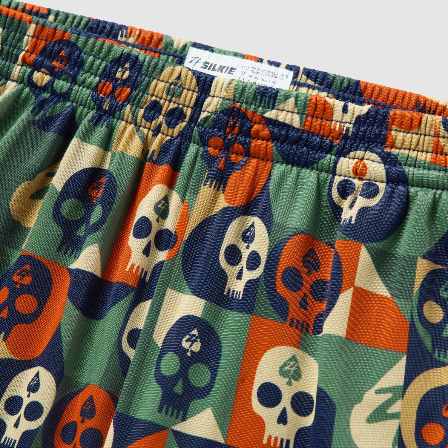 Close-up of lightweight fabric with a colorful geometric skull pattern in blue, orange, cream, and green on Geo Skulls Silkies shorts with an elastic waistband.