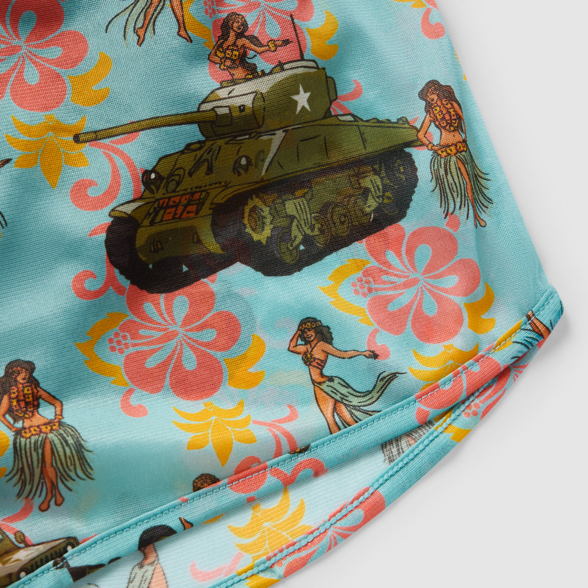 Close-up of light blue fabric from Hula Tank Blue Silkies featuring colorful hibiscus flowers, hula dancers, and a detailed military tank illustration.
