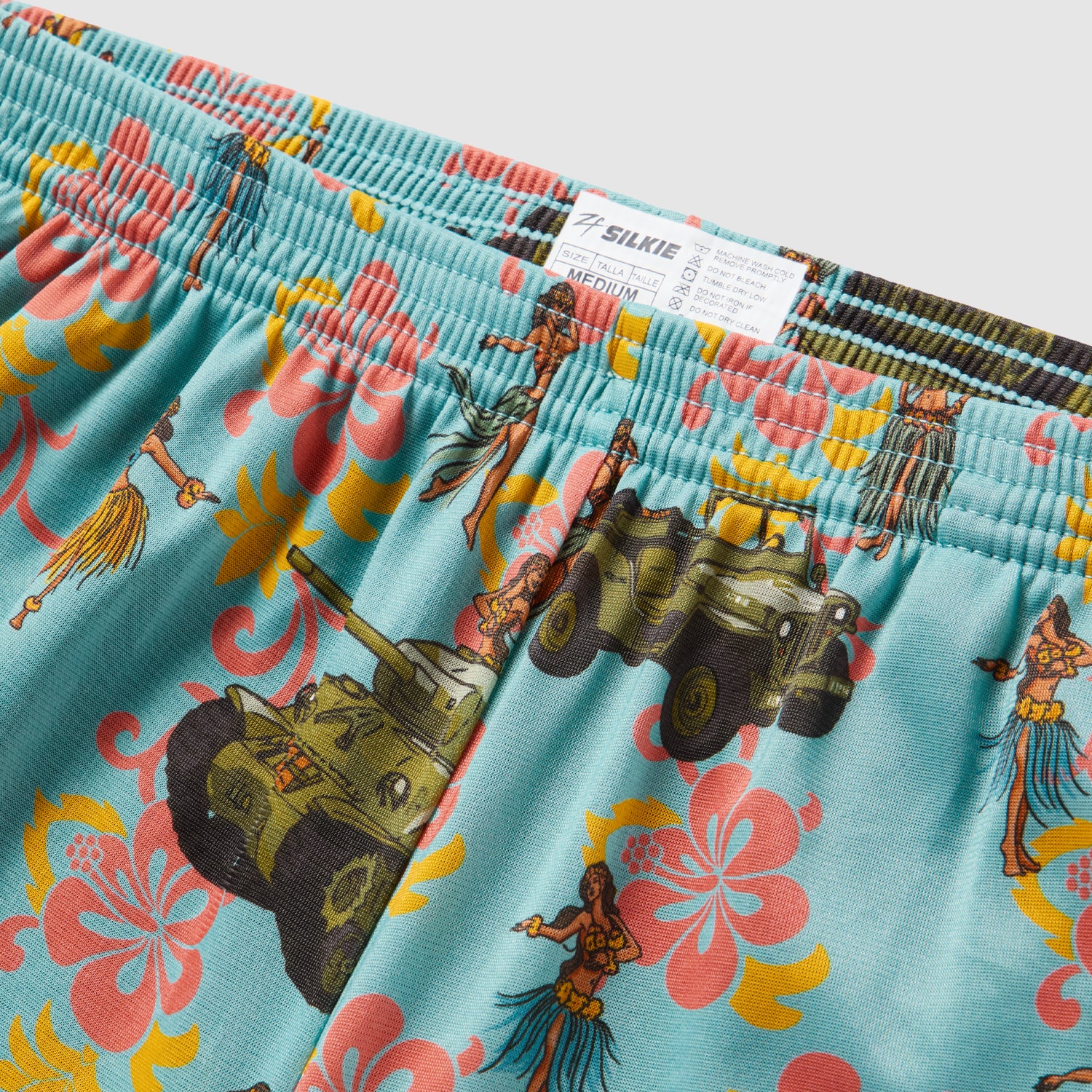 Close-up view of blue patterned fabric featuring colorful floral designs, tropical dancers, and military vehicles, highlighting the waistband and tag of Hula Tank Blue Silkies.