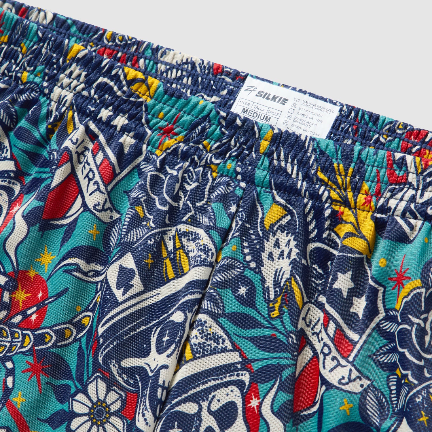 Close-up of colorful patterned fabric featuring skulls, stars, hearts, and banners with the word Liberty on Liberty Skull Silkies shorts with an elastic waistband and size label.