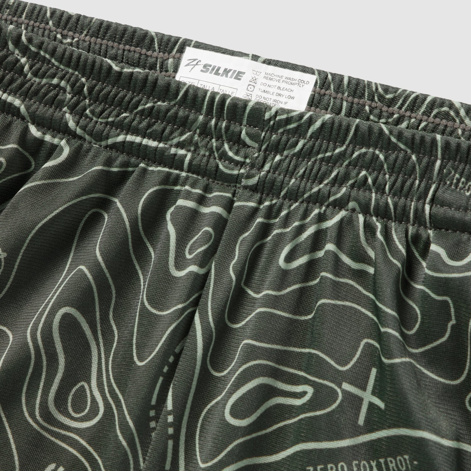 Close-up view of olive green Topographic Silkies featuring a detailed topographic contour line pattern and an elastic waistband with a branded inner label.