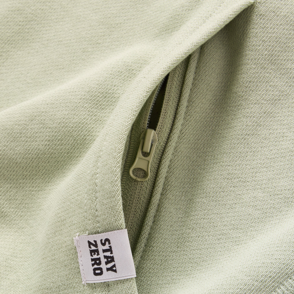 Close-up of a light sage green Smuggler French Terry Crew showing a hidden zipper pocket and a fabric tag with the text "STAY ZERO" on textured fabric.