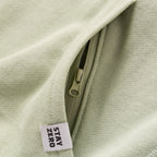 Close-up of a light sage green Smuggler French Terry Crew showing a hidden zipper pocket and a fabric tag with the text "STAY ZERO" on textured fabric.