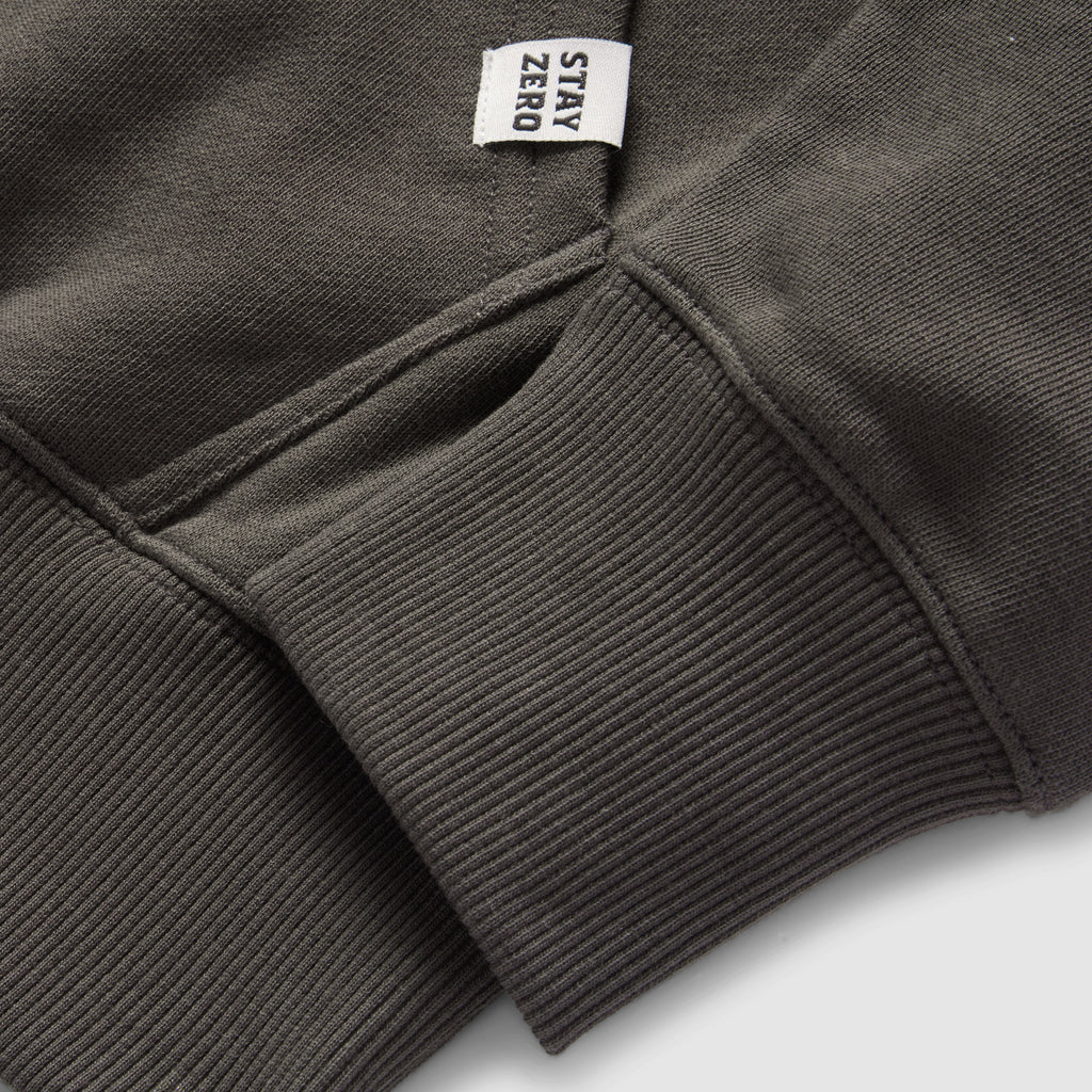 Close-up of a dark gray Smuggler French Terry Crew sleeve showing ribbed cuff detail and a small fabric tag with the text "STAY ZERO" attached near the seam.
