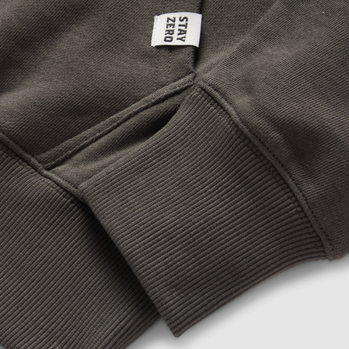 Close-up of a dark gray Smuggler French Terry Crew sleeve showing ribbed cuff detail and a small fabric tag with the text "STAY ZERO" attached near the seam.