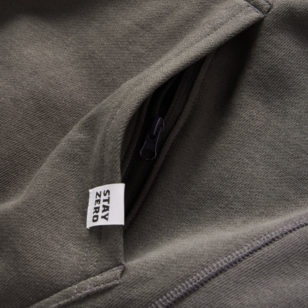 Close-up of an olive green Smuggler French Terry Crew showing a zippered pocket detail and a fabric tag with the words "STAY ZERO.