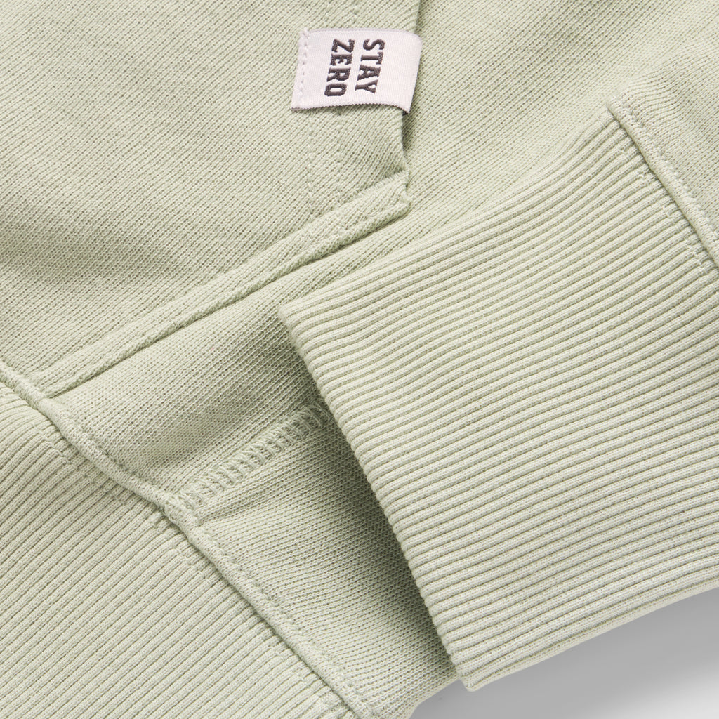 Close-up of the sleeve cuff and pocket seam detail in a soft sage green fabric of the Smuggler French Terry Hoodie with a small white tag that says Stay Zero.