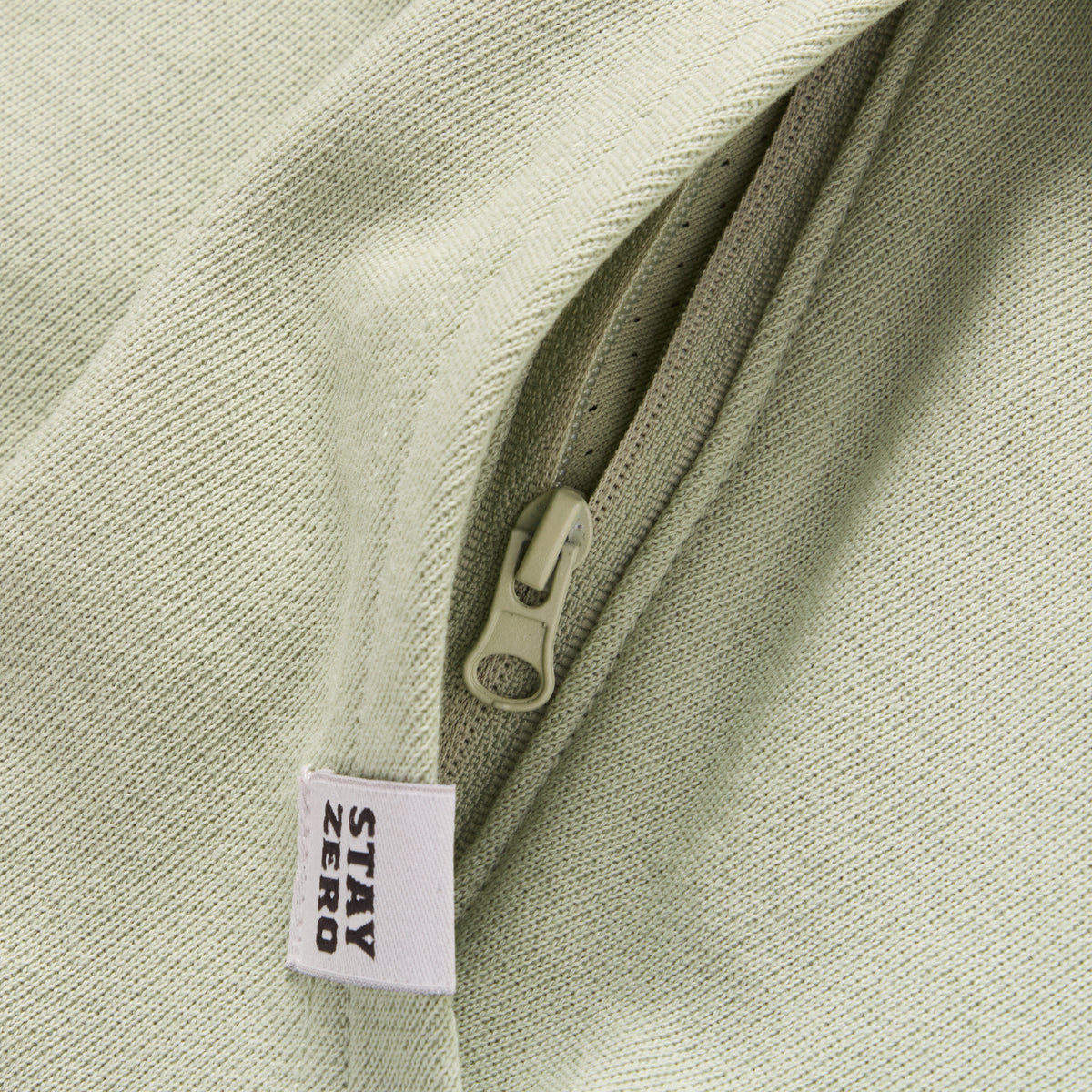 Close-up of a light sage green Smuggler French Terry Hoodie showing the textured fabric, zipper detail, and a "STAY ZERO" fabric tag.