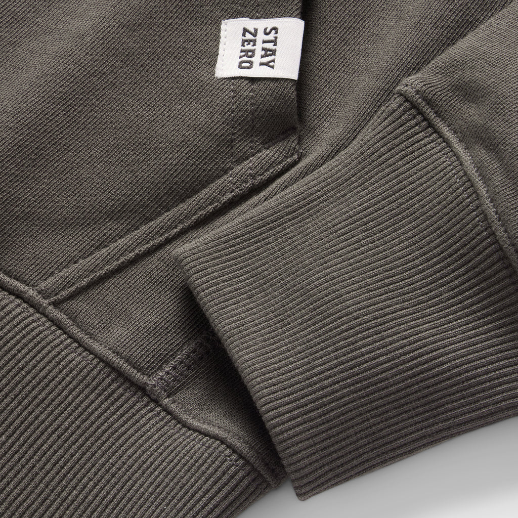 Close-up detail of a dark gray Smuggler French Terry Hoodie showing the ribbed cuff, pocket stitching, and a small white fabric tag with "STAY ZERO" text.