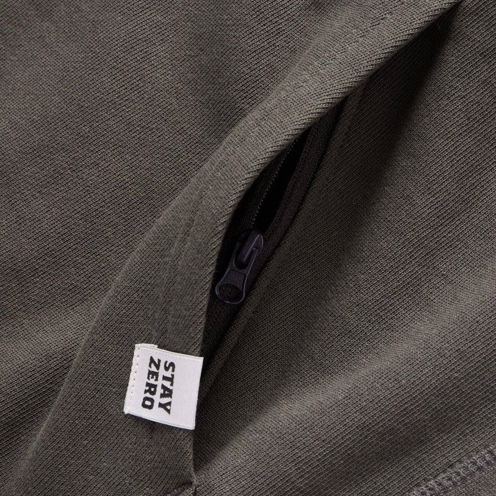 Close-up of an olive green Smuggler French Terry Hoodie featuring a black zipper pocket and a white fabric tag with the text "STAY ZERO" sewn into the seam.