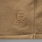 Close-up of tan fabric with embroidered skull and spade logo detail on the Smuggler French Terry Jog Shorts - 6" Inseam showing textured stitching and hem.