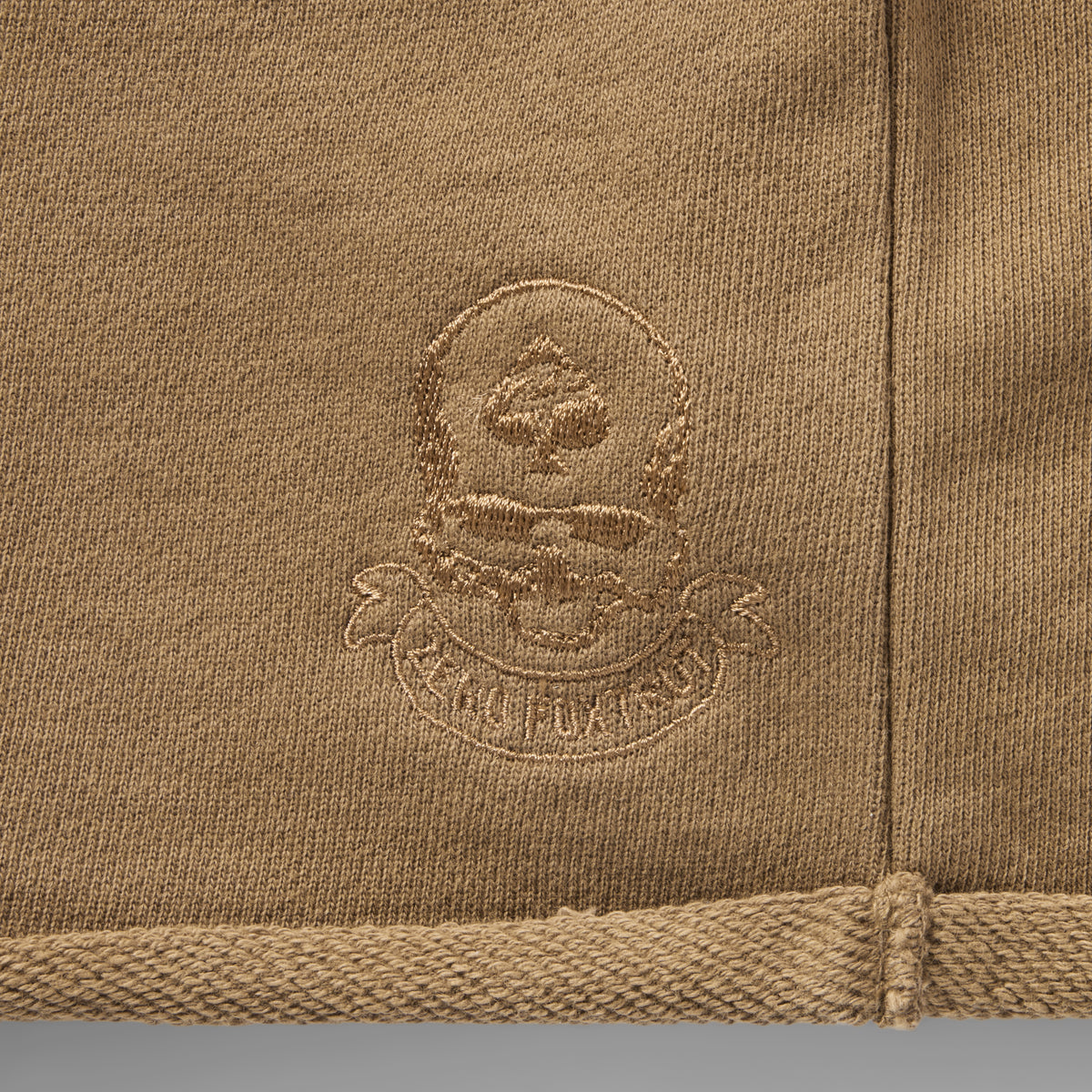 Close-up of tan fabric with embroidered skull and spade logo detail on the Smuggler French Terry Jog Shorts - 6" Inseam showing textured stitching and hem.