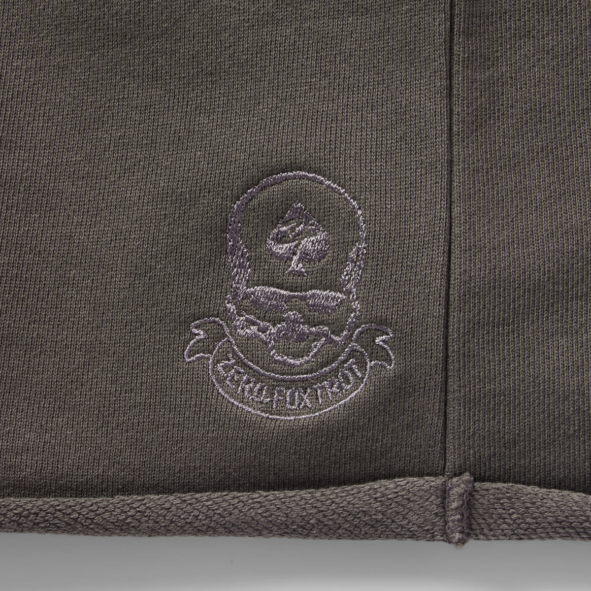 Close-up of embroidered logo detailing on dark fabric of Smuggler French Terry Jog Shorts - 6" Inseam, showcasing texture and stitching quality.