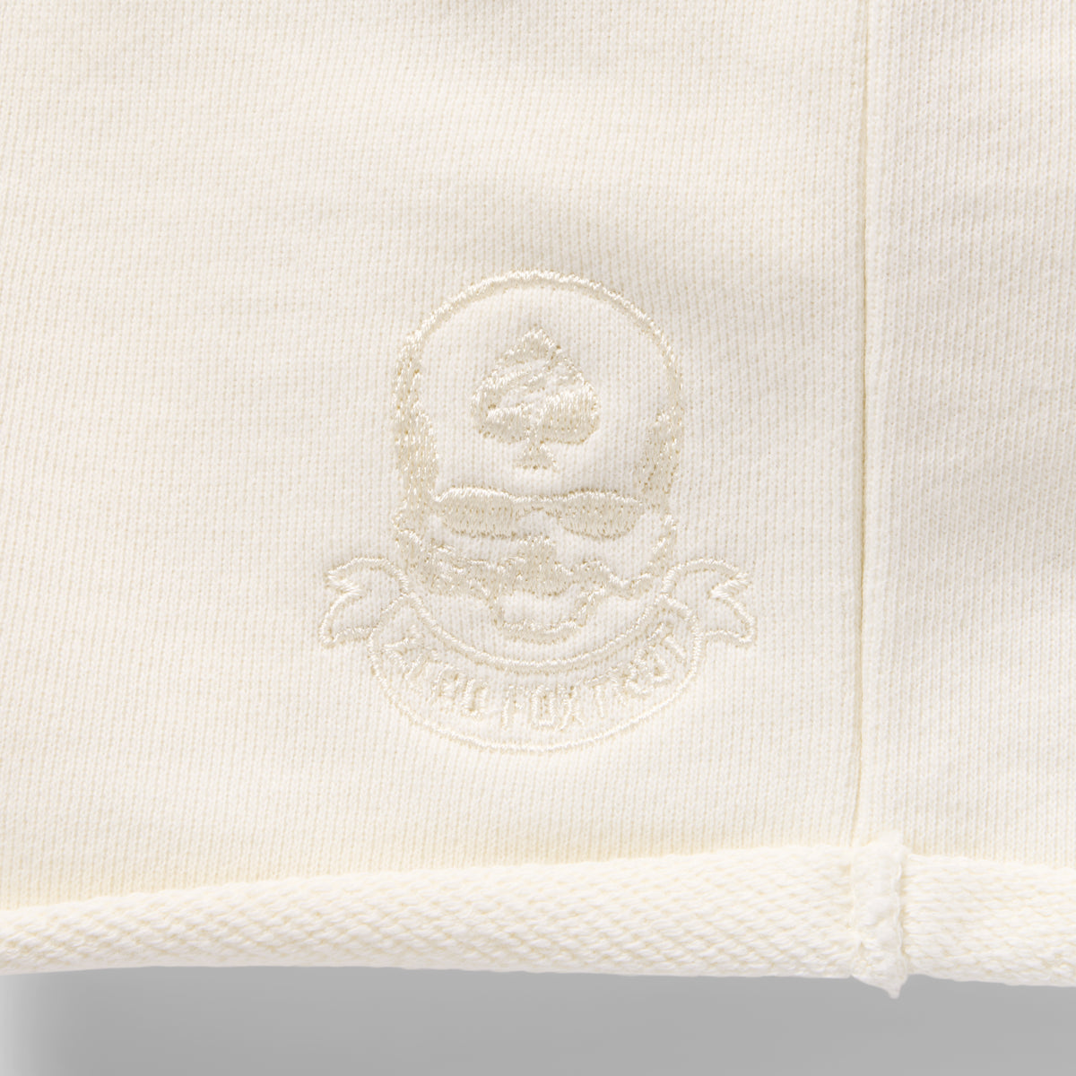 Close-up view of the embroidered logo detail on the light-colored fabric of the Smuggler French Terry Jog Shorts - 6" Inseam.