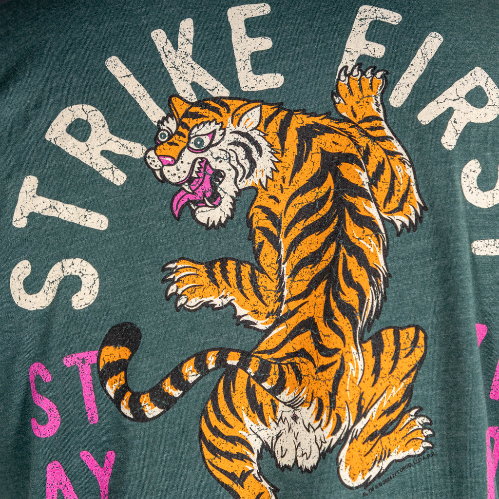 Close-up of a green Strike Hard Tee showing a fierce orange and black tiger graphic with claws extended and the words "STRIKE FIRST" printed above in bold, distressed white letters.