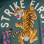 Close-up of a green Strike Hard Tee showing a fierce orange and black tiger graphic with claws extended and the words "STRIKE FIRST" printed above in bold, distressed white letters.