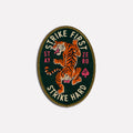 Strike Hard Patch embroidered oval patch featuring a fierce orange tiger on a dark green background with bold cream and red lettering and gold edging