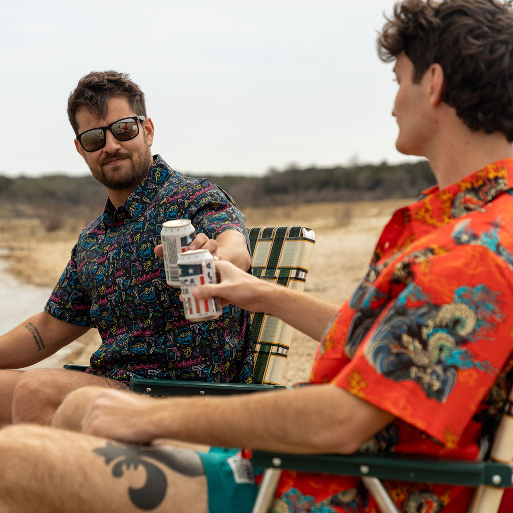 Surf Shirts (Last Chance) featuring two men in colorful patterned shirts relaxing in outdoor chairs, with one offering a canned drink to the other