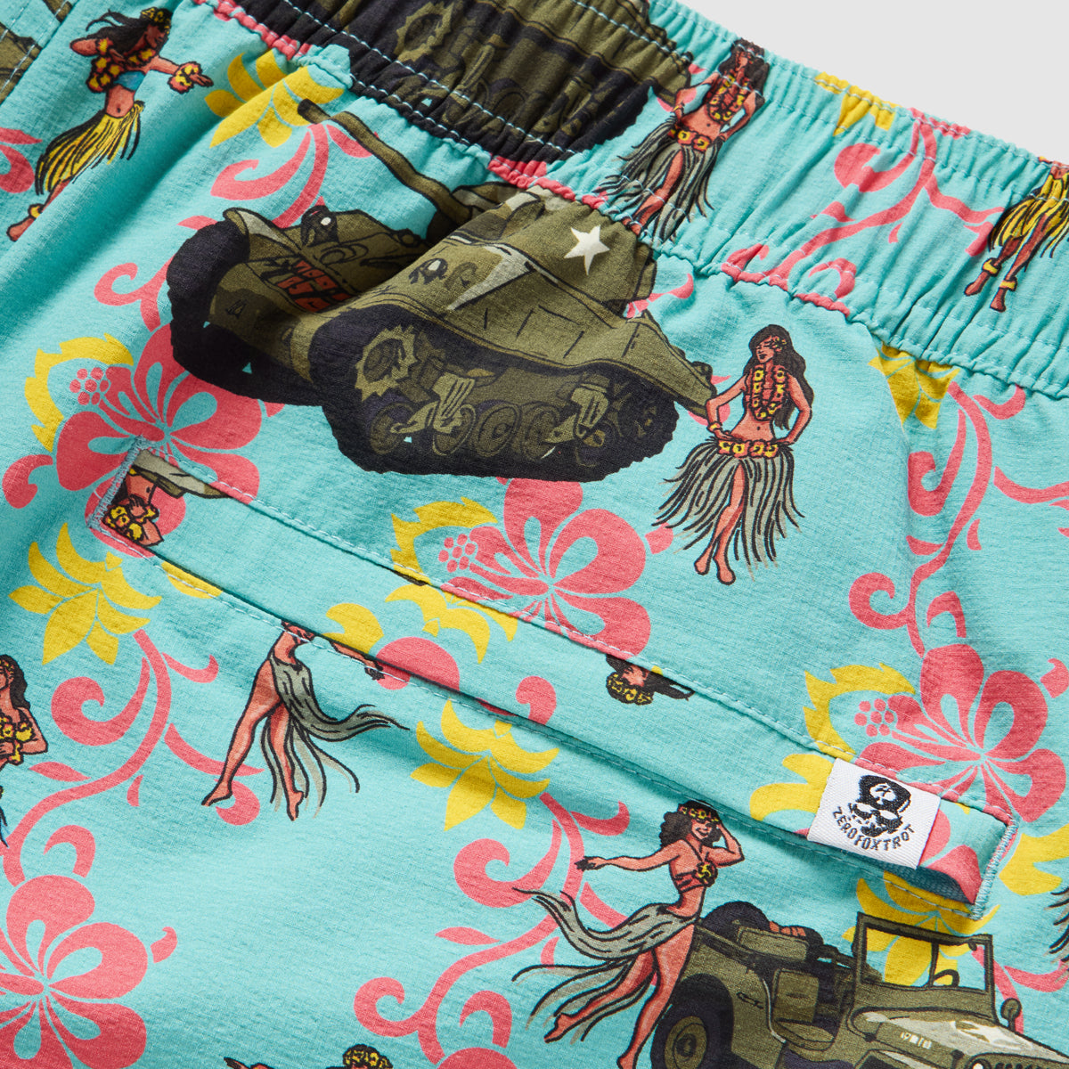 Close-up of blue Swim Trunks featuring a colorful tropical floral pattern with vintage military tanks and hula dancer illustrations, elastic waistband, and a small logo tag on the back pocket.