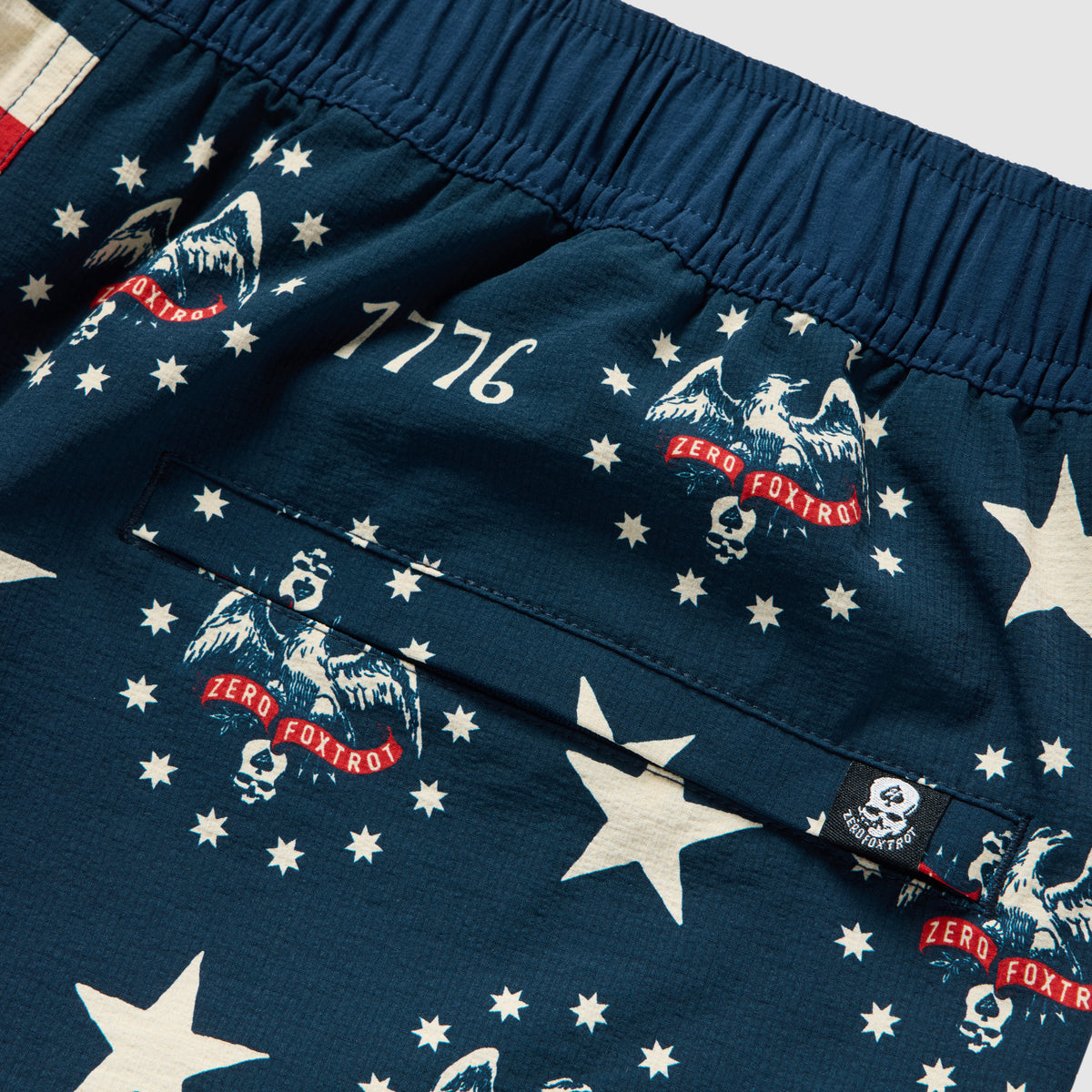 Close-up of navy blue Swim Trunks featuring white stars, the number 7776, and eagle and skull graphics with red "Zero Foxtrot" text on the fabric near a back pocket.