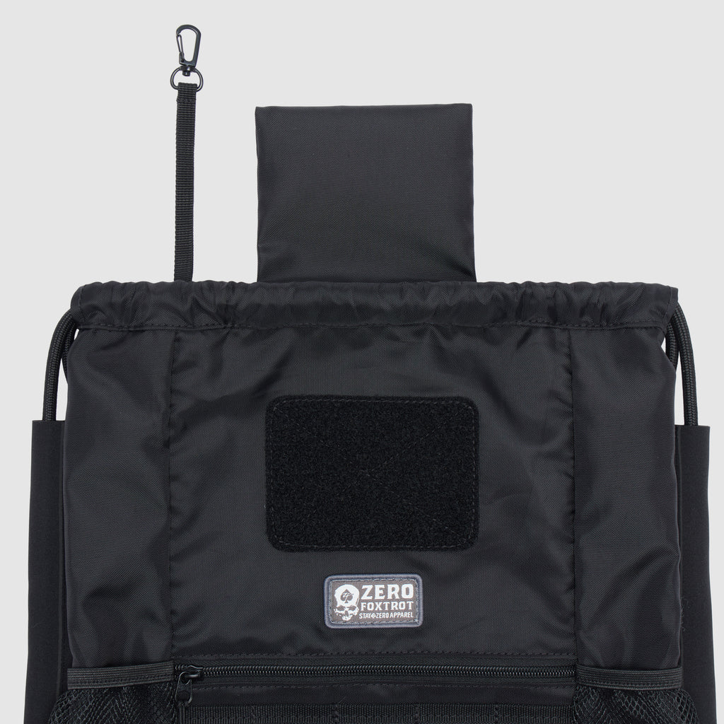 Black TAC Drawstring Pack with an open top, featuring a black hook strap attachment, front Velcro patch area, and a zippered mesh pocket at the bottom.