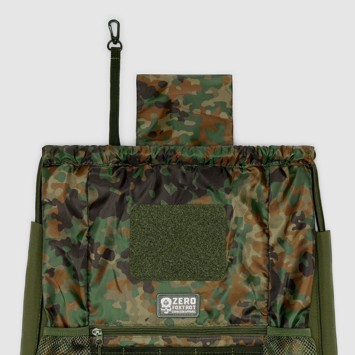 TAC Drawstring Pack in camouflage pattern featuring a top flap, front mesh pocket with zipper, and a small clip attached to a strap for secure closure or hanging.