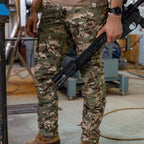Tac Pants in a camouflage pattern with multiple pocket details, worn by a person holding a rifle in an industrial workshop