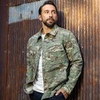 Man wearing a camouflage patterned Chore Jacket over a white shirt, standing against a rustic corrugated metal wall with hands in pockets.
