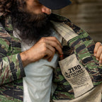 Man wearing a camouflage Chore Jacket with the inner lining exposed, showing a printed pocket label while holding a small red object inside the jacket pocket.