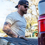 Man wearing sunglasses and a backwards cap, dressed in a light-colored Cotton Skivvy Tee, standing by the open tailgate of a truck outdoors on a sunny day.