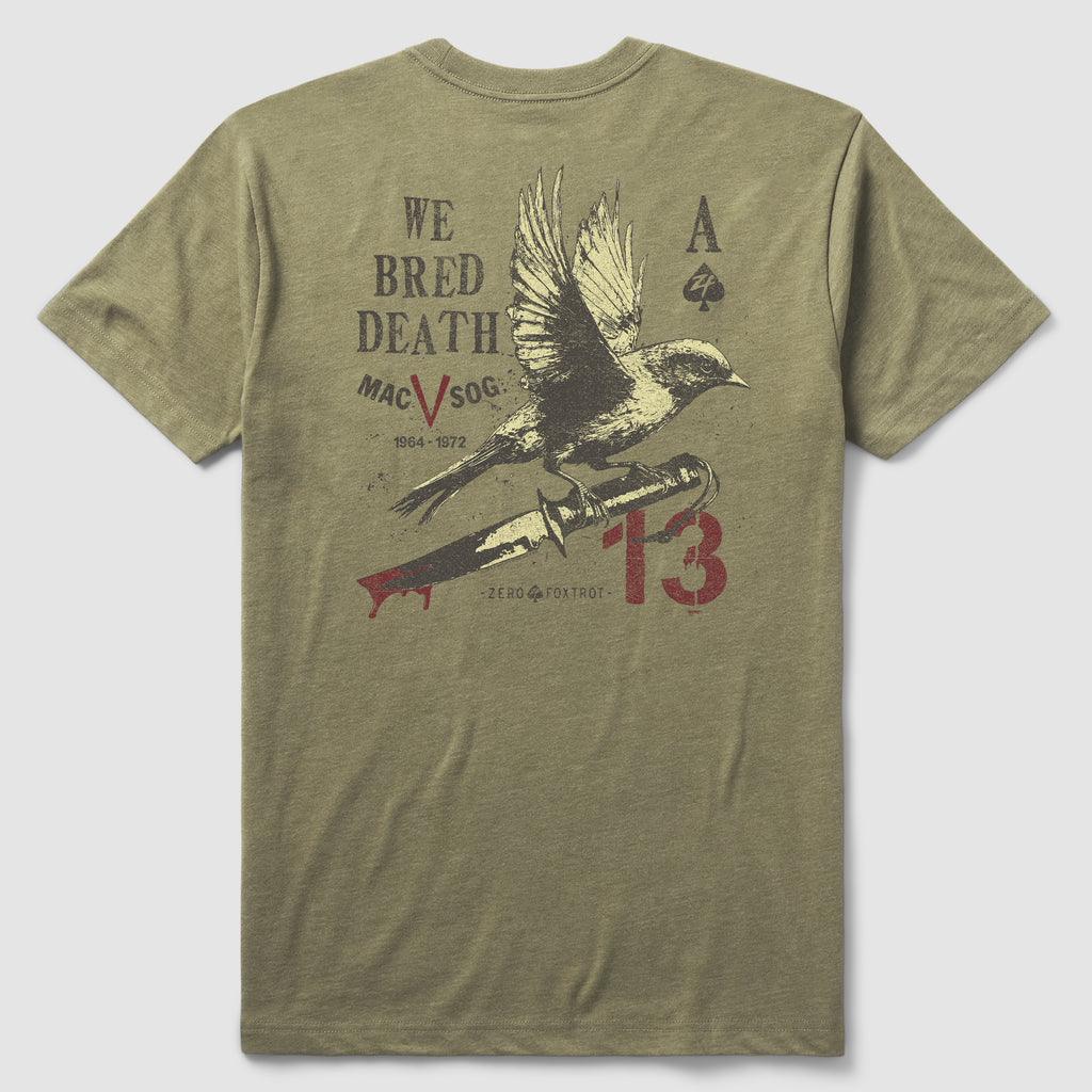 Back view of an olive green We Bred Death Tee featuring a graphic design with a bird perched on a bleeding knife, accompanied by text and symbols including "We Bred Death," "Mac V Soc," and the number 13.