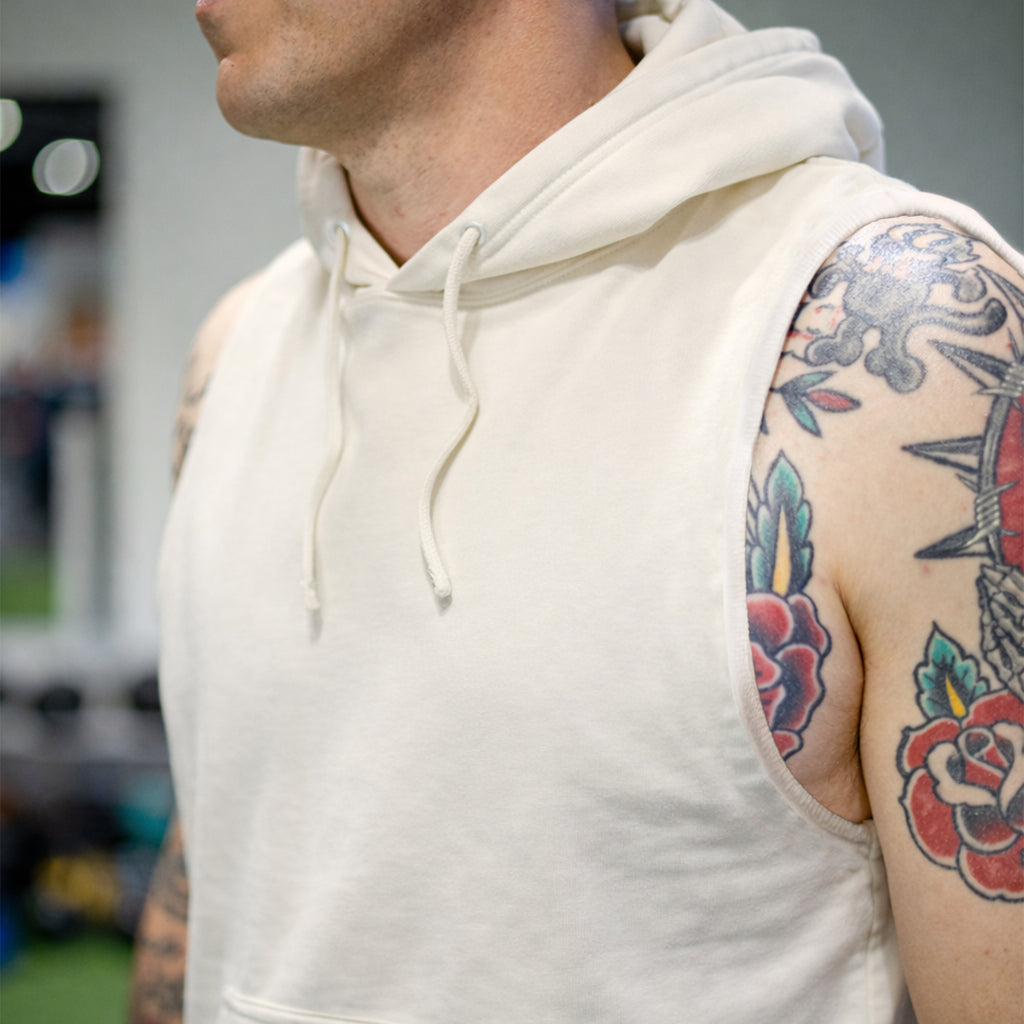 Close-up of a man wearing a sleeveless cream-colored Smuggler French Terry Muscle Hoodie with a drawstring hood, showcasing tattooed arms and a soft textured fabric.