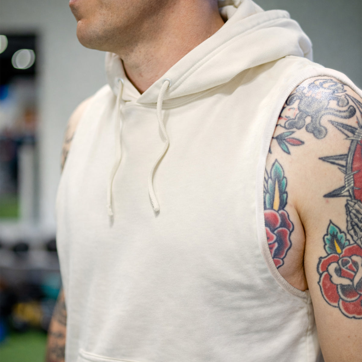 Close-up of a man wearing a sleeveless cream-colored Smuggler French Terry Muscle Hoodie with a drawstring hood, showcasing tattooed arms and a soft textured fabric.