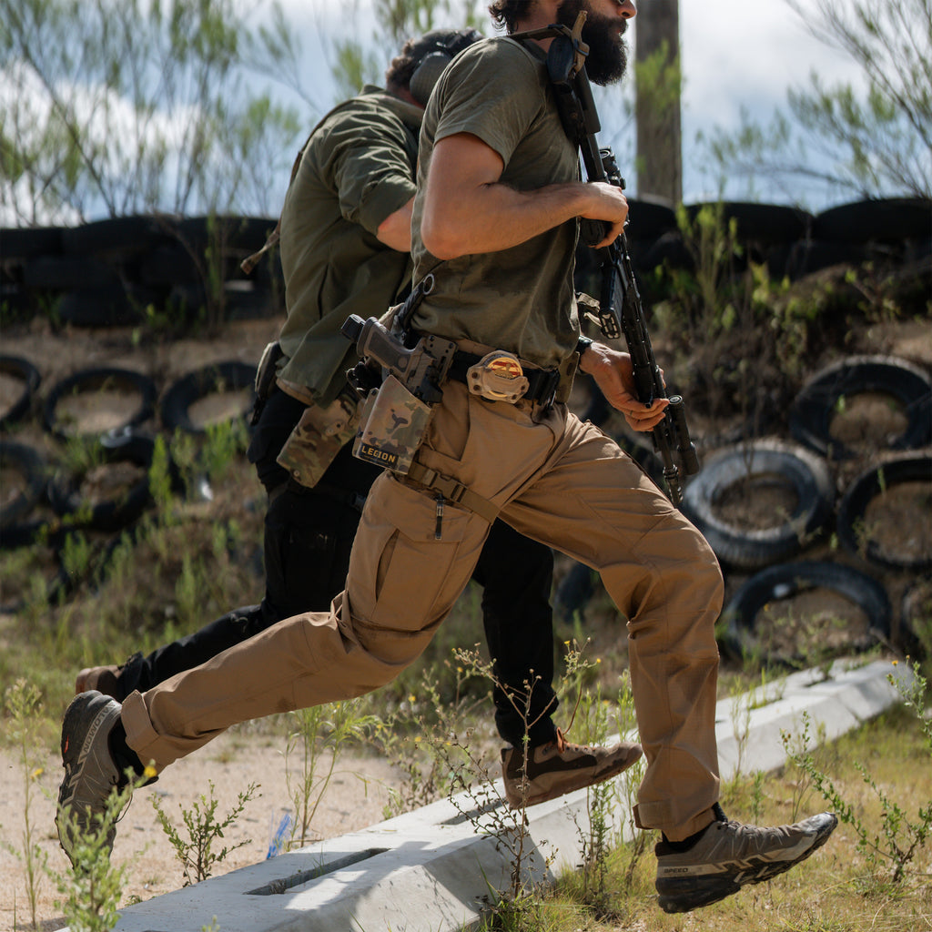 Tac Pants in tan worn by a man running outdoors with tactical gear and cargo pockets visible in a rugged training environment