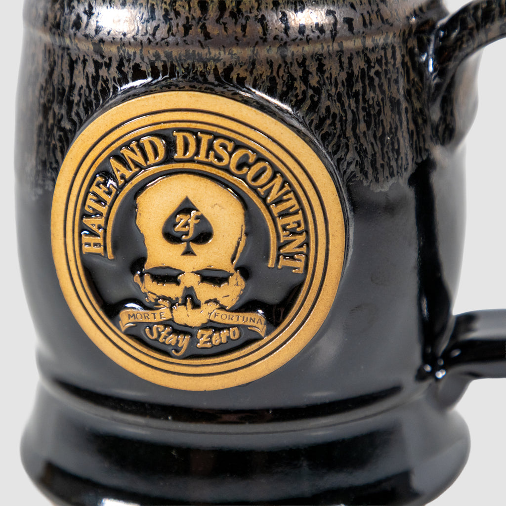 Close-up of a black ceramic Zero Dark Mug featuring a gold emblem with a skull, spade symbol, and the words "Hate and Discontent" and "Stay Zero" on the front.