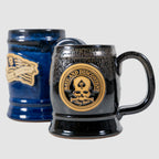Two ceramic Zero Dark Mugs side by side, one in glossy black and the other in deep blue, each featuring a raised emblem with a skull and text design.