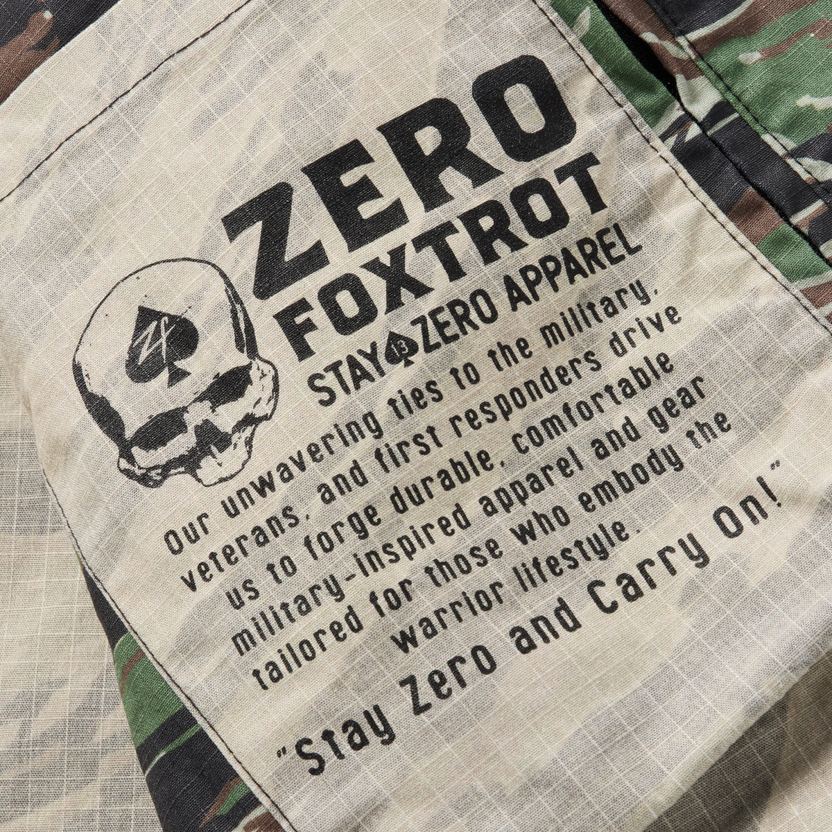 Close-up of a camouflage Chore Jacket featuring a stitched patch with a skull graphic and bold text describing the brand's military-inspired, durable, and comfortable apparel ethos.