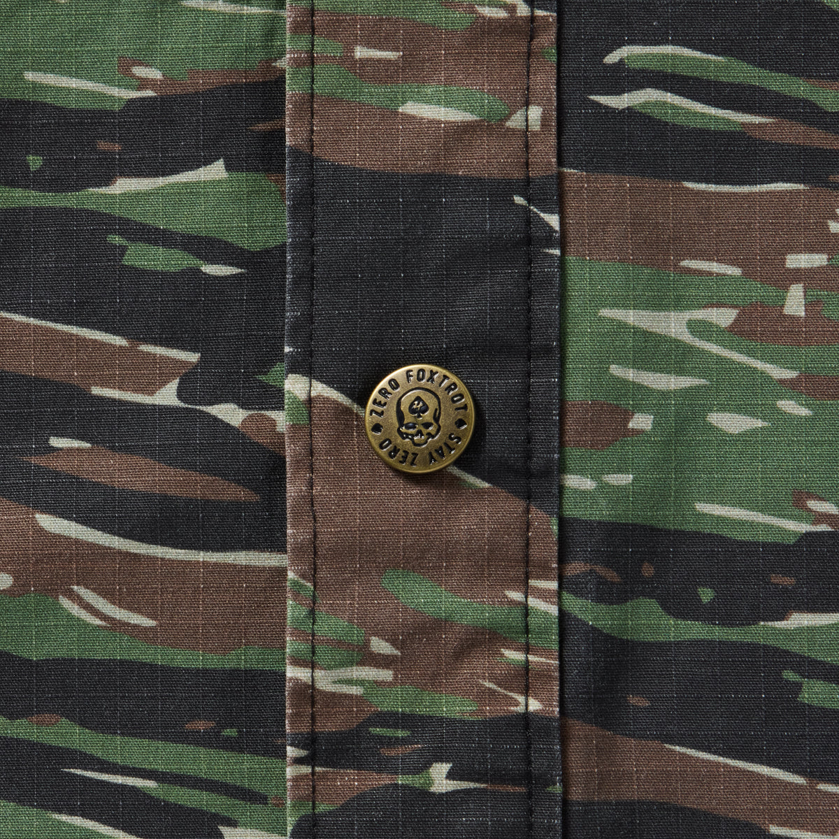 Close-up of a camouflage patterned fabric with a brass snap button featuring a skull and the text "Zero Foxtrot" on the Chore Jacket.