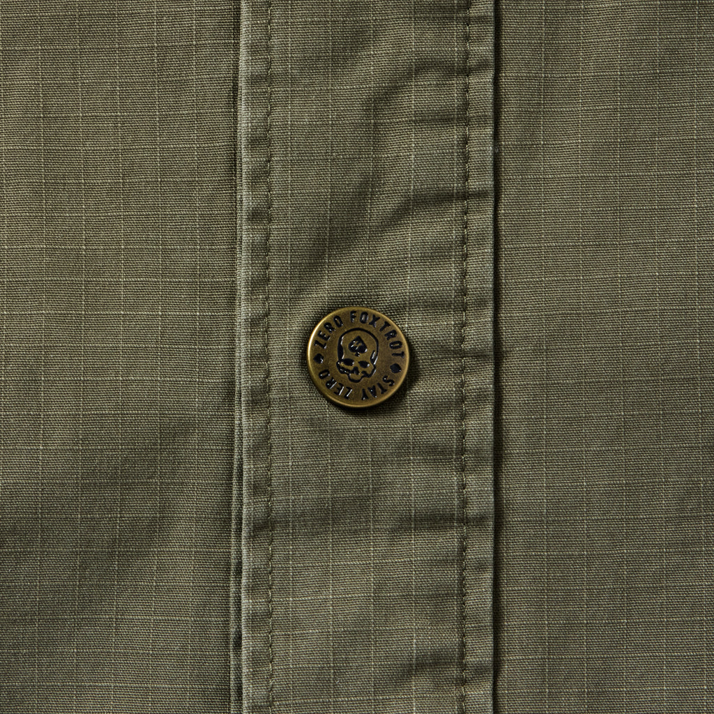 Close-up of a dark olive Chore Jacket showing textured fabric with grid pattern and a detailed metal button featuring engraved text and a skull design.