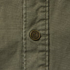 Close-up of a dark olive Chore Jacket showing textured fabric with grid pattern and a detailed metal button featuring engraved text and a skull design.