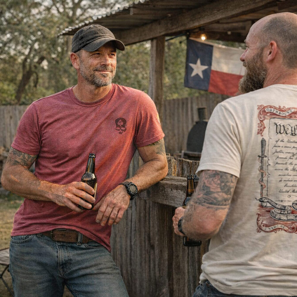 Two men wearing casual t-shirts, one in a red shirt and the other in a beige shirt with a printed design, standing outdoors near a rustic fence with a Texas flag in the background, both holding beer bottles, showcasing the ZF Core Tee 250th Birthday 3-Pack.