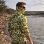 Man wearing a green camouflage Performance Sun Hoodie with an extended neck gaiter, standing by a lake with a rocky shoreline and tree-covered hills in the background.