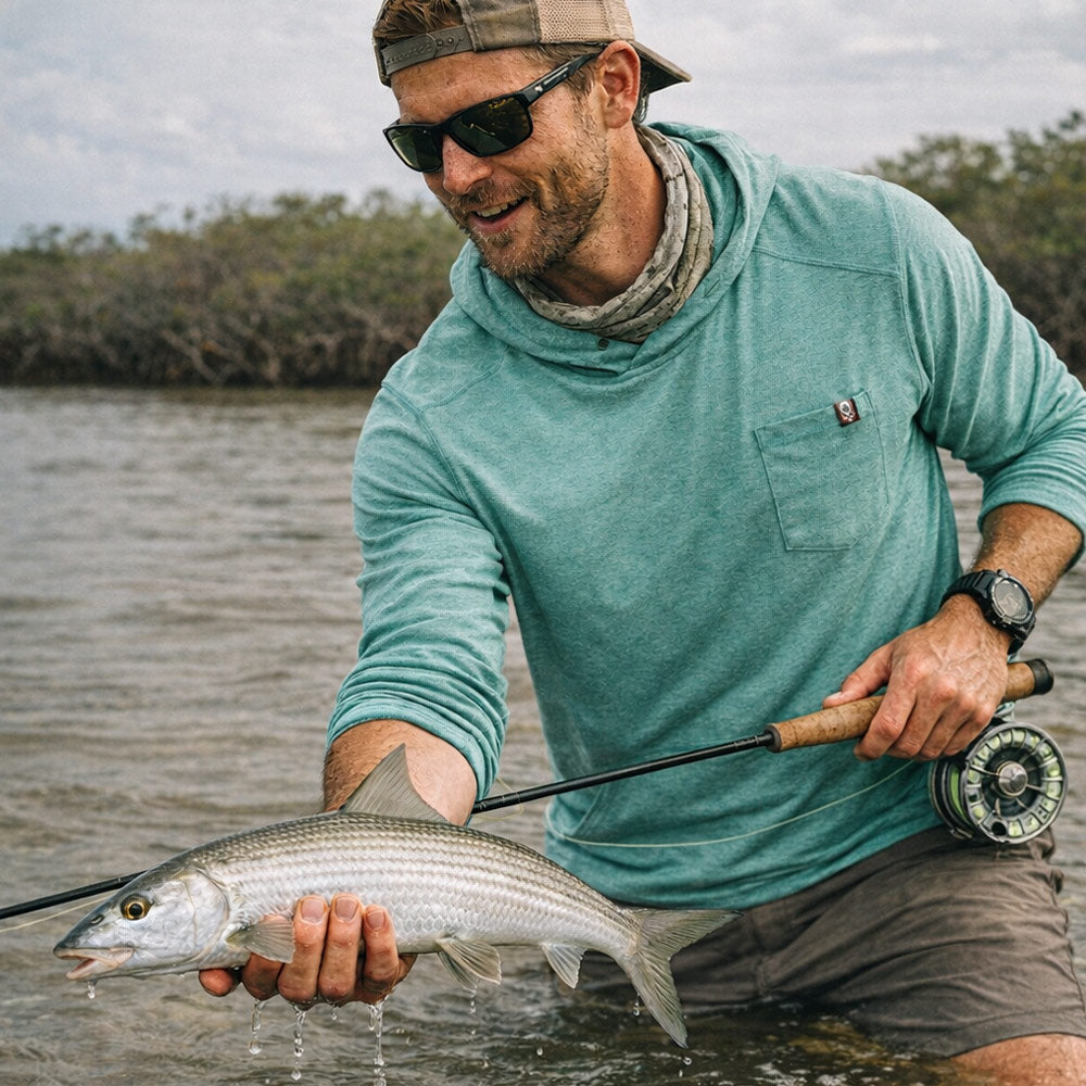 Man wearing a teal Performance Sun Hoodie while fishing and holding a freshly caught fish near the water's edge on a cloudy day.