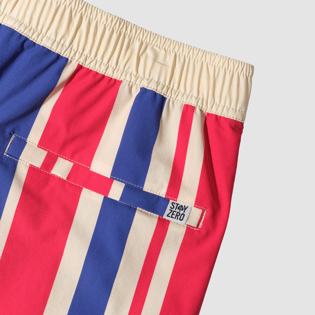 Close-up view of swim trunks with red, blue, and beige vertical stripes, featuring a beige elastic waistband and a back pocket with a small black and white label.
