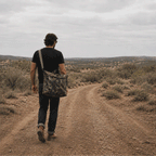 ZF Flyers Helmet Bag carried over the shoulder by a person walking along a rugged desert trail with dry hills and shrubs in the background