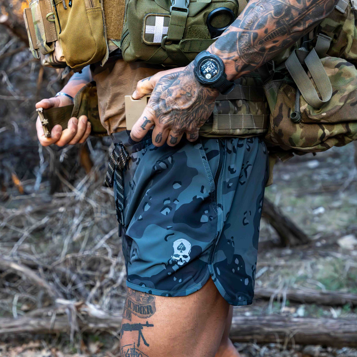 Close-up of a person wearing dark camo patterned Zero Shorts - 5" Inseam outdoors, showing detailed tattoos and tactical gear in a rugged natural setting.
