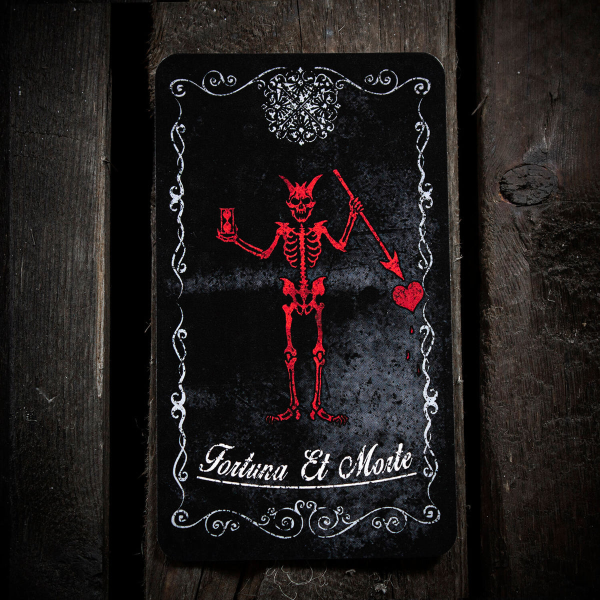 Fortune Tarot Card featuring a red skeleton holding a sword and pointing to a heart on a black card with ornate white borders on dark wood