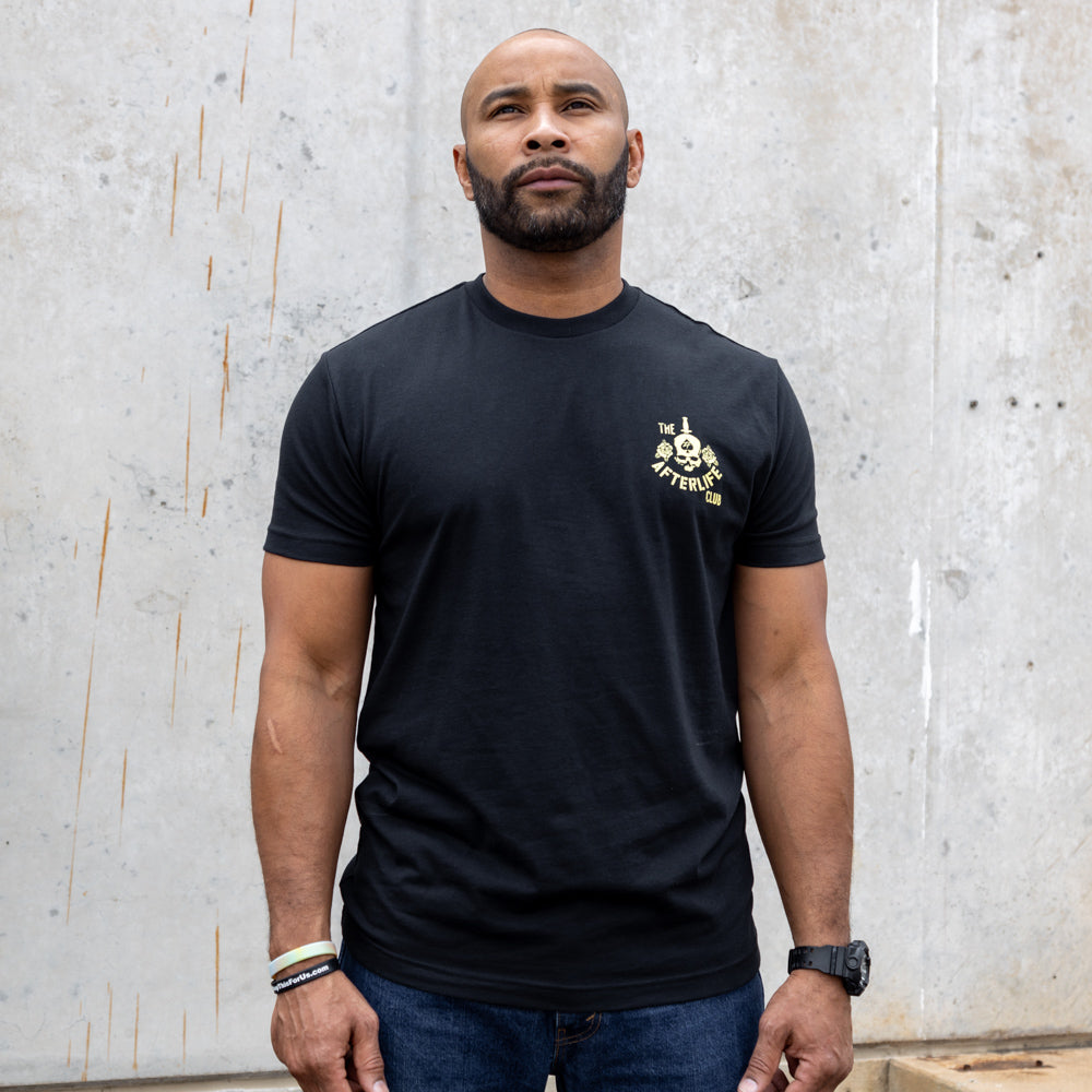 Apple a Day Tee black short sleeve shirt worn by a man standing against a textured concrete wall, featuring a small graphic on the left chest